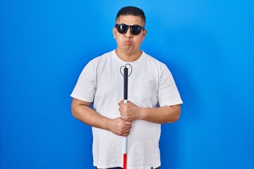 Hispanic young blind man holding cane puffing cheeks with funny face. mouth inflated with air,...