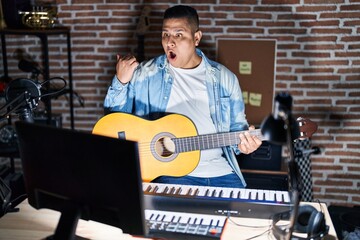Hispanic young man playing classic guitar at music studio surprised pointing with hand finger to...
