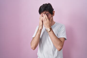 Young hispanic man standing over pink background with sad expression covering face with hands while...