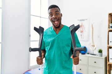 Young african physiotherapist man holding crutches celebrating crazy and amazed for success with...