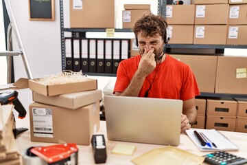 Young hispanic call center agent man working at warehouse smelling something stinky and disgusting,...