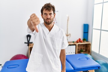 Young handsome physiotherapist man working at pain recovery clinic looking unhappy and angry...