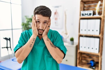 Young physiotherapist man working at pain recovery clinic tired hands covering face, depression and...