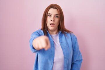 Young hispanic woman with red hair standing over pink background pointing displeased and frustrated...