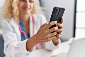 Middle age blonde woman wearing doctor uniform using smartphone at clinic