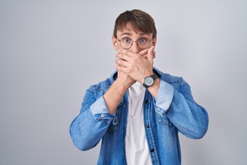 Caucasian blond man standing wearing glasses shocked covering mouth with hands for mistake. secret...