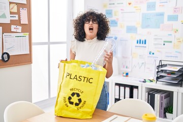 Young middle east woman holding recycling bag with plastic bottles at the office angry and mad...