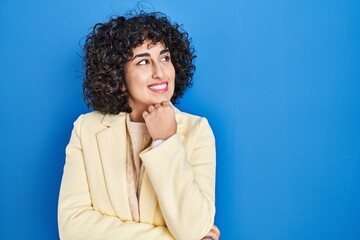 Fototapeta na wymiar Young brunette woman with curly hair standing over blue background with hand on chin thinking about question, pensive expression. smiling and thoughtful face. doubt concept.