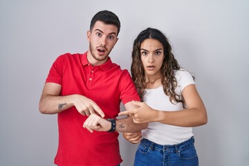 Young hispanic couple standing over isolated background in hurry pointing to watch time,...