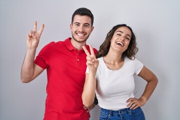 Young hispanic couple standing over isolated background smiling looking to the camera showing fingers doing victory sign. number two.