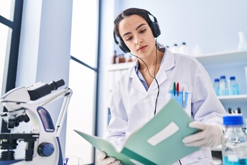Young hispanic woman wearing scientist uniform listening to music reading book at laboratory