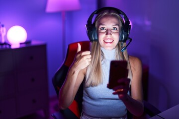 Young caucasian woman playing video games with smartphone smiling happy and positive, thumb up doing excellent and approval sign