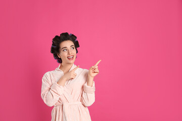 Happy young woman in bathrobe with hair curlers on pink background, space for text