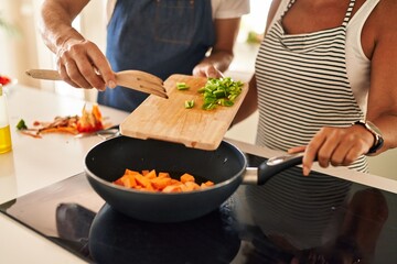 Middle age hispanic couple pouring food on frying pan at kitchen