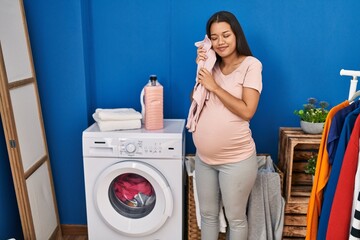 Young latin woman pregnant holding baby clothes at laundry room