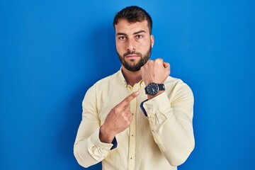 Handsome hispanic man standing over blue background in hurry pointing to watch time, impatience, looking at the camera with relaxed expression