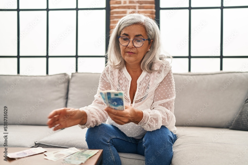 Sticker Middle age woman counting sloty banknotes sitting on sofa at home