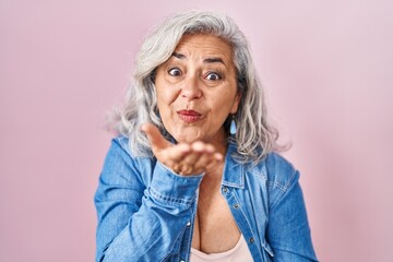 Middle age woman with grey hair standing over pink background looking at the camera blowing a kiss...