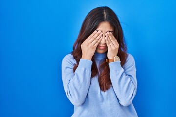 Hispanic young woman standing over blue background rubbing eyes for fatigue and headache, sleepy...