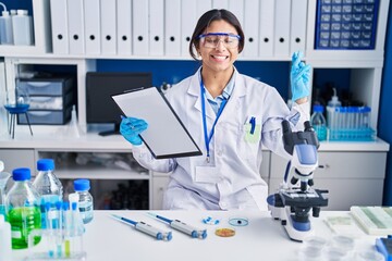 Hispanic young woman working at scientist laboratory gesturing finger crossed smiling with hope and eyes closed. luck and superstitious concept.