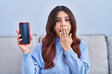 Hispanic young woman holding smartphone showing blank screen covering mouth with hand, shocked and...