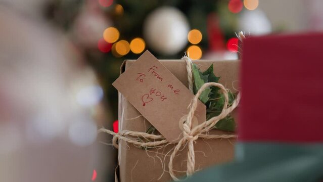 Close up of gift box with tag FROM ME TO YOU against Christmas tree lights bokeh. Eco friendly gift wrapping