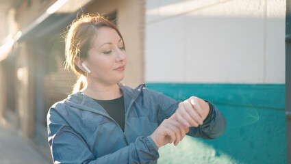 Young redhead woman wearing sportswear listening to music looking stopwatch at street