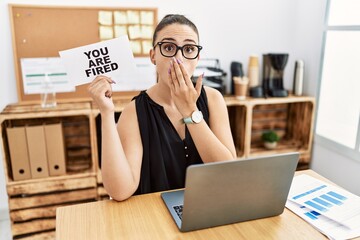 Young brunette woman holding you are fired banner at the office covering mouth with hand, shocked...