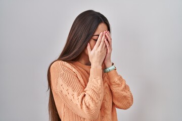 Young brunette woman standing over white background with sad expression covering face with hands...