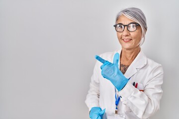 Fototapeta premium Middle age woman with grey hair wearing scientist robe cheerful with a smile of face pointing with hand and finger up to the side with happy and natural expression on face