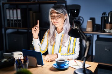 Middle age woman with grey hair working at the office at night showing and pointing up with finger number one while smiling confident and happy.
