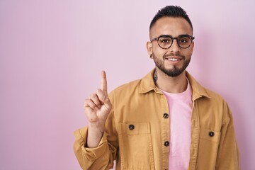 Young hispanic man standing over pink background showing and pointing up with finger number one while smiling confident and happy.