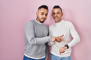 Homosexual couple standing over pink background in hurry pointing to watch time, impatience, upset...