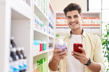 Young hispanic man customer using smartphone holding gel bottle at pharmacy