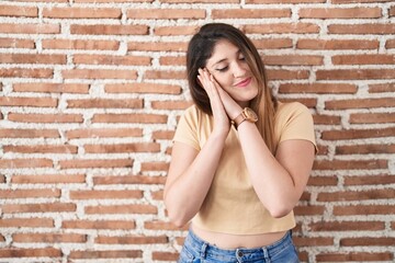 Young brunette woman standing over bricks wall sleeping tired dreaming and posing with hands together while smiling with closed eyes.