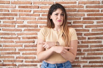 Young brunette woman standing over bricks wall pointing to both sides with fingers, different direction disagree