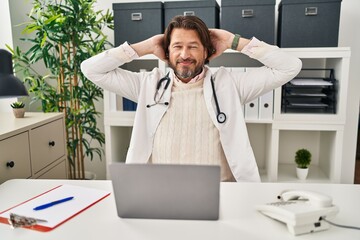 Handsome middle age doctor man working at the clinic relaxing and stretching, arms and hands behind head and neck smiling happy