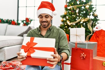 Young hispanic man unboxing gift sitting on floor by christmas tree at home
