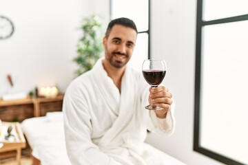 Young hispanic man wearing bathrobe toasting with wine at beauty center