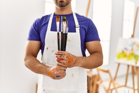 Young hispanic man holding paintbrushes at art studio