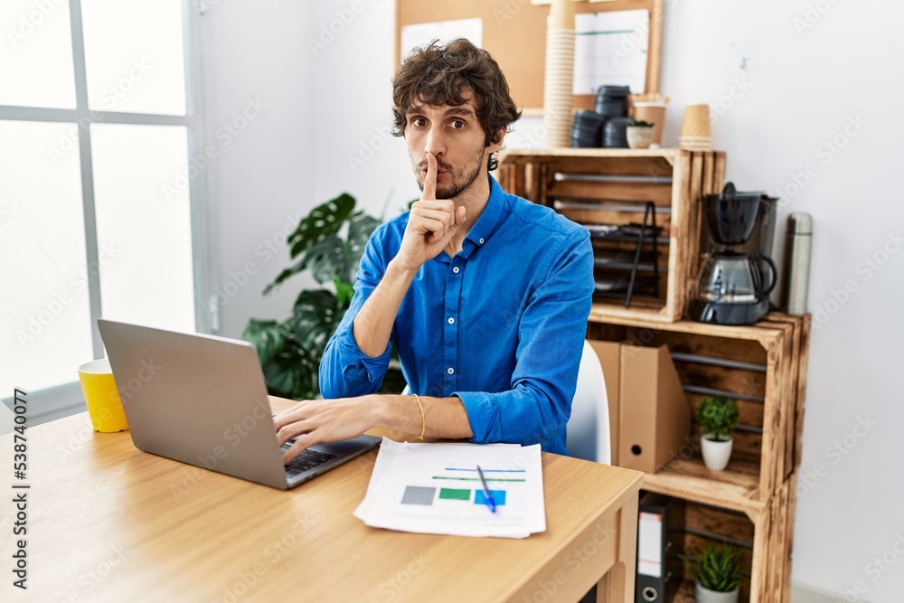 Sticker Young hispanic man with beard working at the office using computer laptop asking to be quiet with finger on lips. silence and secret concept.