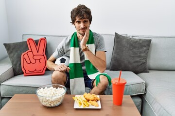 Young hispanic man football hooligan cheering game at home touching mouth with hand with painful expression because of toothache or dental illness on teeth. dentist concept.