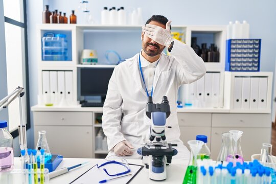 Young Hispanic Man With Beard Working At Scientist Laboratory Smiling And Laughing With Hand On Face Covering Eyes For Surprise. Blind Concept.