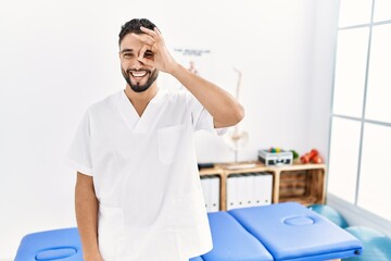 Young handsome man with beard working at pain recovery clinic doing ok gesture with hand smiling, eye looking through fingers with happy face.