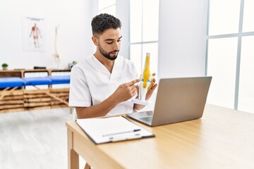 Young arab man wearing physiotherapist uniform having video call holding anatomical model of knee...