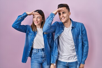 Obraz premium Young hispanic couple standing over pink background very happy and smiling looking far away with hand over head. searching concept.