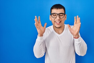 Naklejka premium Young arab man wearing casual white shirt and glasses celebrating mad and crazy for success with arms raised and closed eyes screaming excited. winner concept