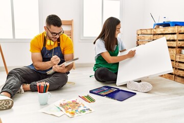 Young hispanic couple smiling happy drawing sitting on the floor at art studio.