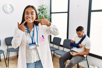 Obraz premium Young asian doctor woman at waiting room with a man with a broken arm smiling cheerful showing and pointing with fingers teeth and mouth. dental health concept.