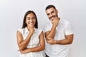 Young interracial couple standing together in love over isolated background looking confident at the camera smiling with crossed arms and hand raised on chin. thinking positive.